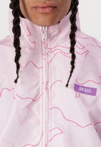 Light pink jacket with a wavy pink pattern, full zip closure, high collar, and a purple branded label on the front left.