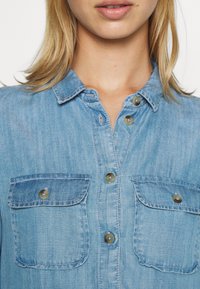 Vaaleansininen farkkupaita, jossa on napitettava kaulus, kaksi rintataskua ja pyöreät napit. Kankaan pinta on pehmeä ja teksturoitu.