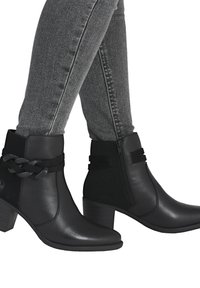 Bottines noires à cheville avec une finition en cuir lisse, dotées d'une sangle tressée, d'une fermeture éclair sur le côté et d'un talon empilé. Portées avec un jean skinny gris.