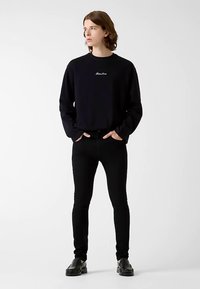 Sweat-shirt noir à manches longues avec logo, associé à un jean noir ajusté et des chaussures noires. Design simple, texture lisse et coupe près du corps.