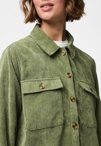 Zielona kurtka koszulowa z sztruksu z ostrym kołnierzem, dwoma zapinanymi na guziki kieszeniami na piersiach oraz zapięciem na guziki z przodu. Charakteryzuje się pionową, prążkowaną teksturą.