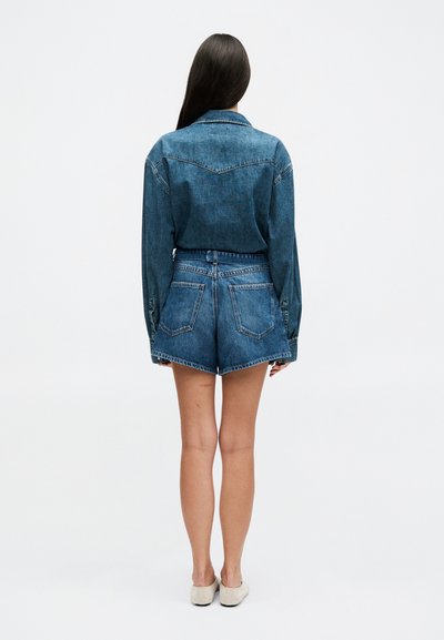 Γυναίκα με μακριά σκούρα μαλλιά, φοράει μπλε τζιν σακάκι και τζιν σόρτς με ψηλή μέση, όρθια ξυπόλυτη και γυρισμένη με την πλάτη.
