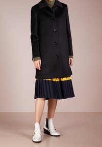 Cappotto di lana nero con cinque bottoni e colletto, abbinato a una gonna plissettata blu navy con un accento giallo senape, e stivaletti bianchi alla caviglia con borchie.