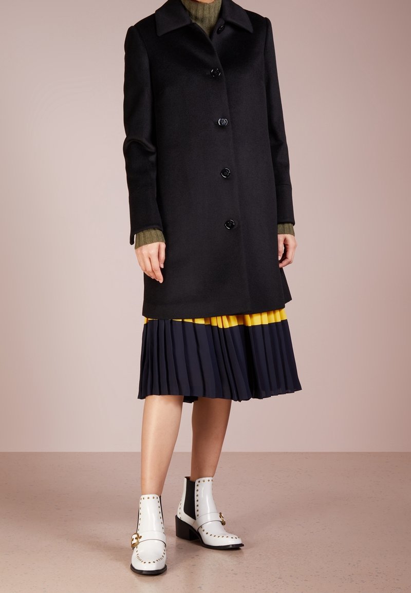 Cappotto di lana nero con cinque bottoni e colletto, abbinato a una gonna plissettata blu navy con un accento giallo senape, e stivaletti bianchi alla caviglia con borchie.
