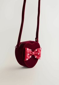 Czerwono-welwetowa torebka w kształcie serca na krzyż, z zamknięciem na zipper, ozdobiona różową satynową kokardą z cyrkoniami. W zestawie regulowany pasek.