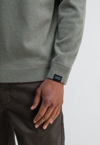 Hellgrüner Pullover aus strukturiertem Stoff, mit gerippten Bündchen und einem gestickten schwarzen Etikett mit der Aufschrift "Butcher of Blue." Getragen über dunklen Hosen.