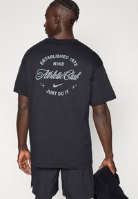 Μαύρο t-shirt Nike με στρογγυλή λαιμόκοψη. Στο πίσω μέρος εμφανίζεται λευκό κείμενο: "Established 1972", "Athletic Club" και "Just Do It." Κοντά μανίκια.