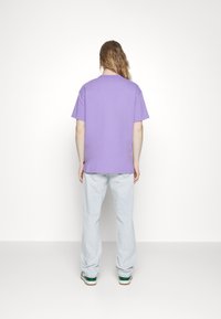 Paarse katoenen T-shirt met korte mouwen en een losse pasvorm, gecombineerd met lichtblauwe jeans en sneakers met groene accenten.