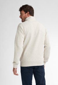 Męski sweter w jasnym beżu, z teksturowanym splotem, wysokim kołnierzem i ściągaczami na rękawach. W połączeniu z ciemnoniebieskimi spodniami.