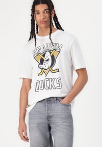 T-shirt blanc à manches courtes présentant un design graphique d'un canard avec des crosses de hockey, le texte "ANAHEIM DUCKS" en lettres noires en gras, associé à un jean gris.