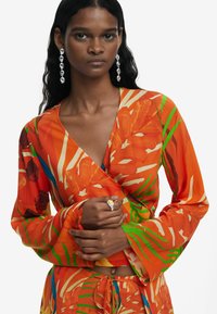 Lys orange indpakningstop med blomster- og bladmønstre i grøn og gul. Lange, svævende ærmer og en V-hals design. Sølv statement øreringe.