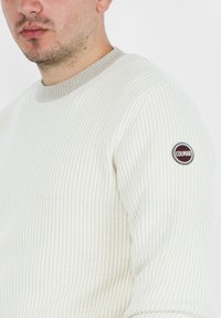Maglione bianco a costine con scollo rotondo e rifiniture beige. Presenta un patch rotondo con il logo sulla manica sinistra. Texture morbida, vestibilità rilassata.