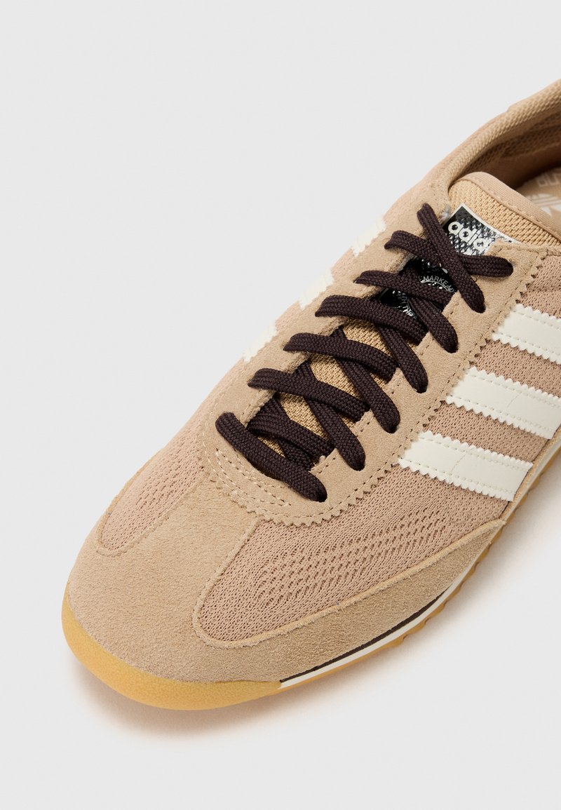 Zapatilla deportiva beige con materiales de ante y malla, rayas blancas y cordones marrones. Punta texturizada y suela de goma para mayor tracción.