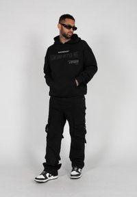 Svart hettegenser med grafisk tekst, paret med svarte cargo-bukser med lommer og stropper. Sko: svart og hvite joggesko.