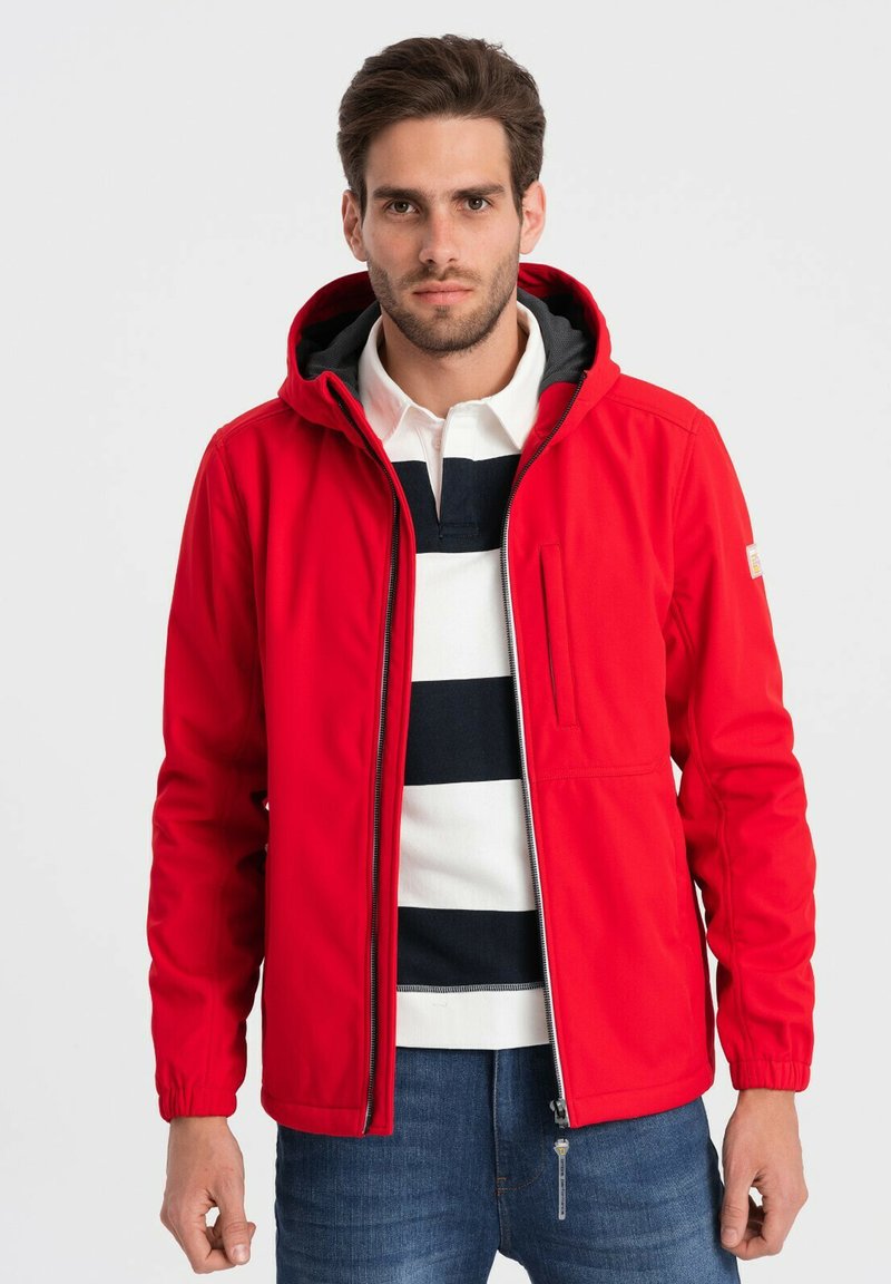 Giacca rossa con zip e cappuccio, caratterizzata da polsini elasticizzati, una tasca laterale con zip e una fodera scura a contrasto. Indossata sopra un polo a righe.