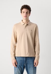Ilgų rankovių smėlio spalvos polo marškiniai su apykakle, turintys mažą logotipą ant krūtinės, pagaminti iš minkštos medžiagos. Dėvimi su mėlynomis džinsinėmis kelnėmis.