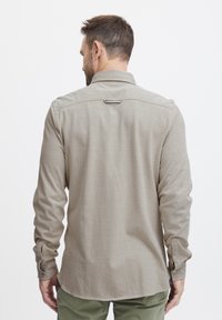 Chemise à manches longues pour homme en tissu beige avec des poignets à bouton, un col rabattu et une petite languette d'accent à l'arrière. Texture lisse et coupe décontractée.