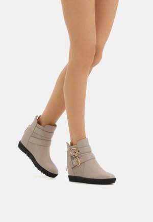 Beige Stiefel mit Knöchelhöhe aus strukturiertem Leder, doppelten Riemenakzenten mit goldenen Schnallen und einer schwarzen Gummiwedge-Sohle für besseren Halt.