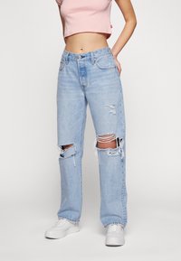 Jeans strappati di colore azzurro chiaro con una vestibilità ampia e orli sfilacciati. Texture di denim visibile, usurati alle ginocchia con un classico design a cinque tasche.