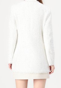 Blazer blanco texturizado con un corte holgado, bordes con flecos y pequeñas motas metálicas. Presenta mangas largas y un corte de longitud media.
