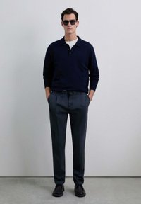 Polo de punto azul marino, camiseta blanca debajo, pantalones grises con cordón, zapatos negros y gafas de sol. Diseño minimalista y ajuste a medida.