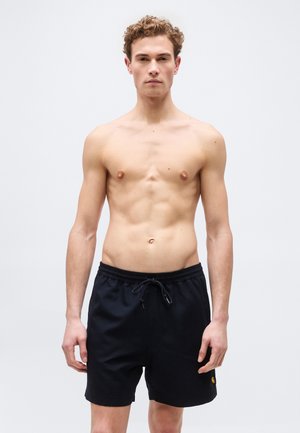 Ung mand med krøllet hår, stående uden skjorte iført sorte shorts med snøre mod en ensartet lys baggrund.