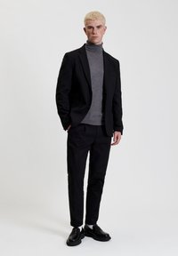 Blazer nero su un dolcevita grigio, abbinato a pantaloni neri. Il modello indossa scarpe nere, ha i capelli biondi corti, in piedi contro uno sfondo semplice.