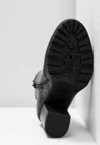 Bottine noire en cuir avec semelle à crampons texturée et talon carré, positionnée semelle vers le haut contre un coin blanc.