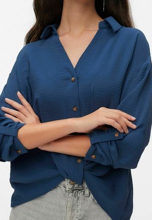 Personne portant une chemise bleue texturée à boutons avec les manches retroussées, les bras croisés et un jean en denim gris clair.
