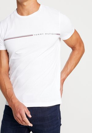 Homme portant un t-shirt blanc à manches courtes avec col rond, rayures horizontales rouges et noires et texte "Tommy Hilfiger", associé à un jean bleu foncé.