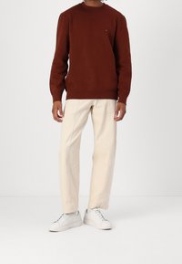 Felpa marrone con scollatura a girocollo, abbinata a pantaloni beige chiari e sneakers bianche. I tessuti sembrano lisci con dettagli minimi.