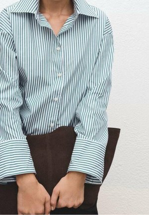 Personne portant une chemise à rayures vertes et blanches avec boutons, tenant une grande pochette marron foncé au niveau de la taille.