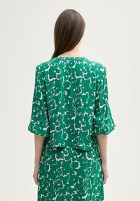 Grüne Bluse mit floralem Muster und Fledermausärmeln, die rosa und weiße Akzente aufweist und eine lockere Passform hat, die am Kragen gerafft ist.