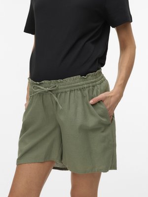 Ženska oblečena v kratke hlače olivno zelene barve z elastičnim pasom in vrvico ter črno kratko majico, roke v žepih hlač, bela ozadje.