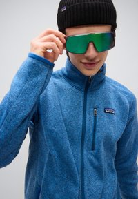 Νεαρός που προσαρμόζει μεγάλα πράσινα αντανακλαστικά γυαλιά ηλίου, φορώντας μπλε fleece Patagonia με φερμουάρ και μαύρο πλεκτό σκουφάκι σε απλό φόντο.