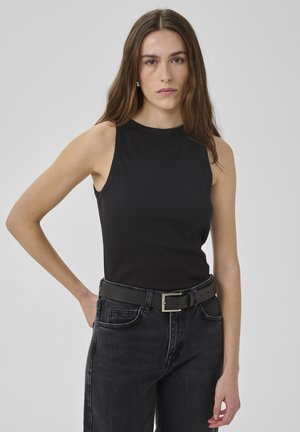Donna con capelli castani lunghi che indossa un top nero senza maniche, cintura nera e jeans scuri, in piedi su uno sfondo grigio uniforme.