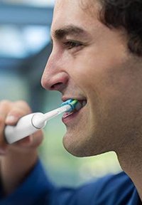Spazzolino elettrico con manico bianco e testina di setole blu, verdi e gialle, tenuto da una persona mentre si lava i denti.
