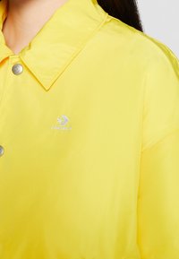 Veste jaune boutonnée avec col, avec le logo Converse blanc brodé sur la poitrine, portée par une personne aux cheveux foncés.