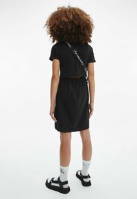 Frau mit lockigem Haar, die ein schwarzes Kleid, weiße Calvin Klein Socken, schwarze Sandalen und eine Umhängetasche mit markiertem Tragegurt trägt, vor weißem Hintergrund stehend.