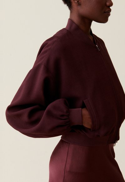 Veste bomber bordeaux avec un col côtelé et des manches bouffantes ; comprend des poches latérales et une texture lisse.