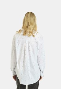 Camicia bianca a maniche lunghe con un motivo testurizzato di piccoli punti metallici. Presenta un colletto button-down e un orlo leggermente curvato.