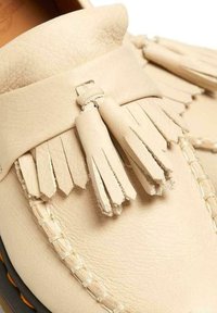 Mocassino in pelle beige con frange decorative, cuciture a contrasto, texture morbida e design della punta arrotondata.