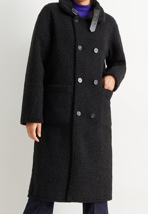 Personne portant un long manteau noir texturé croisé avec de gros boutons, des poches avant et un détail boucle sur le col.