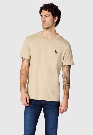 Jonge man met krullend haar, die een beige T-shirt met korte mouwen draagt met een klein palmboomlogo en een blauwe spijkerbroek, kijkt naar links.