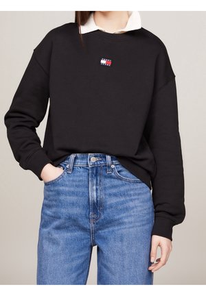 Personne portant un sweat-shirt noir Tommy Jeans sur une chemise blanche à col et un jean bleu taille haute, une main dans la poche.