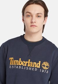 Navigatieblauwe sweatshirt met ribbelkraag, geborduurd "Timberland ESTABLISHED 1973" in geel en grijs, met een accent van het boomlogo.