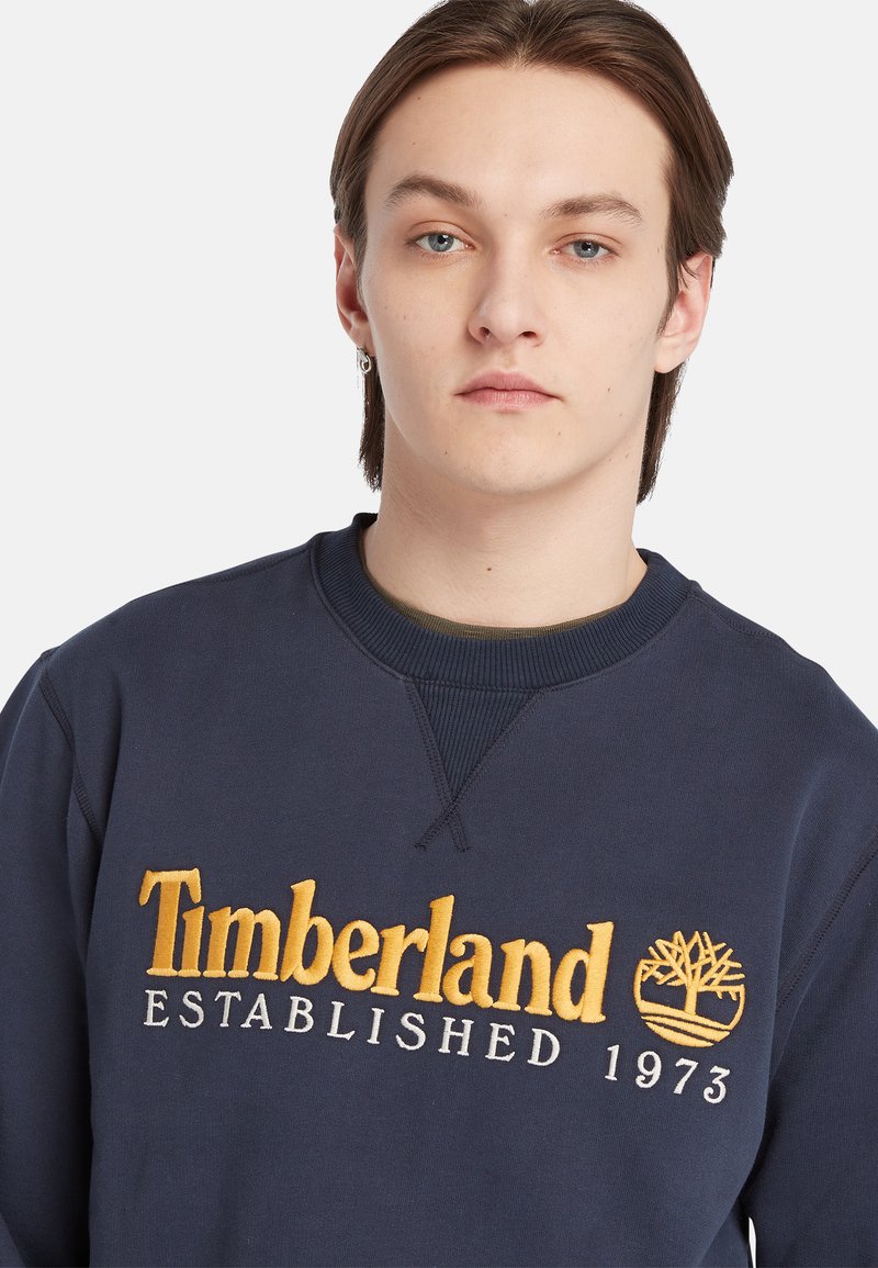 Felpa blu navy con colletto a coste, ricamata con la scritta "Timberland ESTABLISHED 1973" in giallo e grigio, con un logo ad albero come accento.
