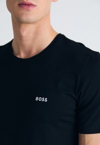 Schwarzes T-Shirt mit kurzen Ärmeln aus glattem Stoff, das auf der linken Brustseite ein weißes gesticktes "BOSS"-Logo trägt.