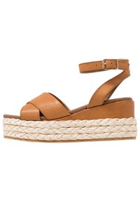Tan leren sandaal met kruisriemontwerp en enkelgesp. Bevat een gevlochten jute platformzool. Soepele textuur met een strakke afwerking.