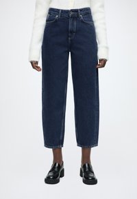 Τζιν σκούρου χρώματος, με ψηλή μέση και ίσια γραμμή, πέντε τσέπες, αντίθετη ραφή και ακατέργαστο τελείωμα. Συνδυασμένο με μαύρα μοκασίνια με πλατφόρμα.